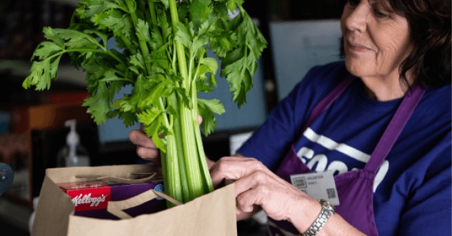 Woman in an apron from Food Bank packing celery into a paper Woolworths shopping bag