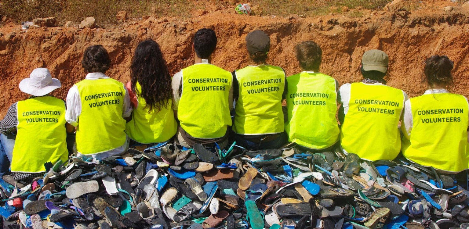 Several volunteers are photographed from behind sat upon a pile of discarded shoes. They are wearing bright yellow vests with the words 'Conservation Volunteers' written across the back.