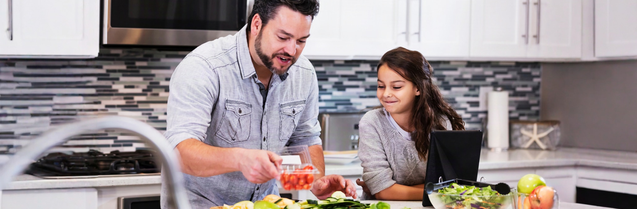 Father is preparing a salad in a light and airy kitchen watched by young daughter who is looking at an iPad