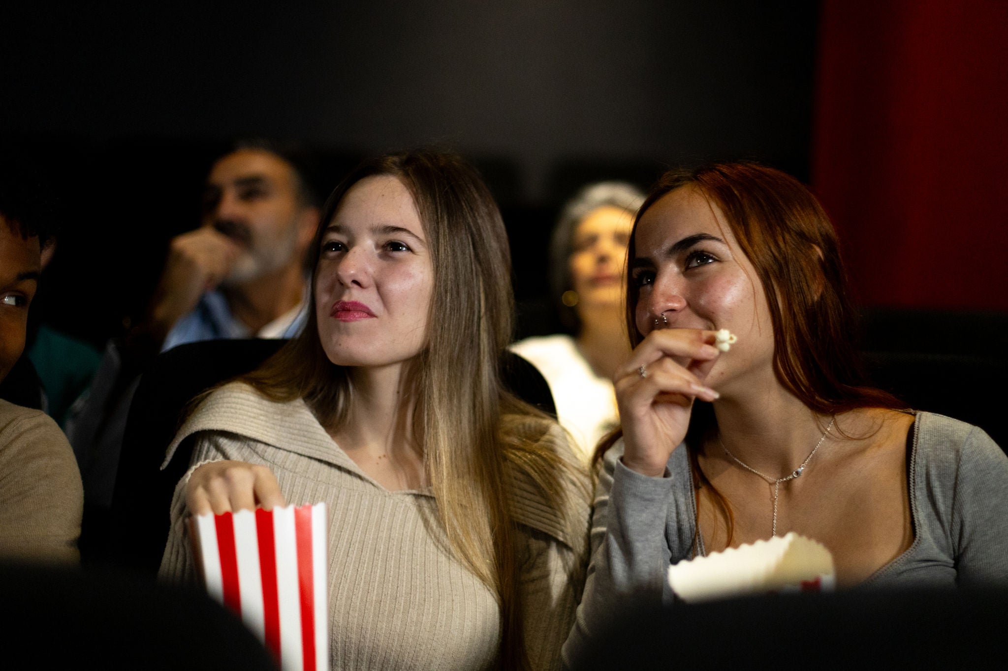 Two young women in a dark movie theatre eating popcorn out of red and white striped boxes