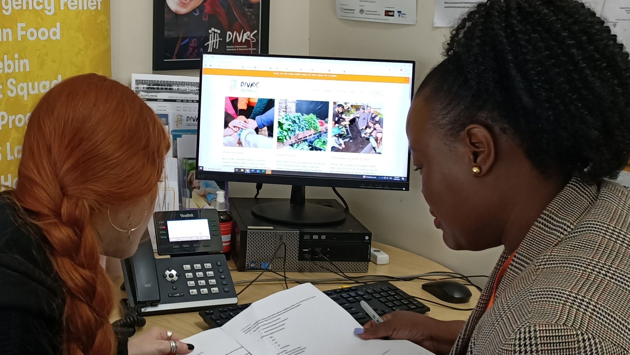 Two women pictured from behind sit at a desk checking some papers together. They are sat in the corner of the room and the walls are covered in various posters and notices from DIVRS. There is a computer monitor and phone on the desk in front of them.
