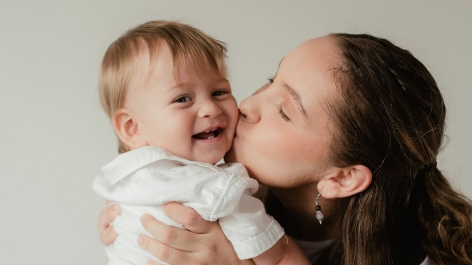 A young mother sits in a chair with closed eyes and kisses her baby on the cheek. The baby is strawberry blonde, smiling, looks towards the camera.