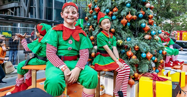 Smiling children dressed as elves sitting on the Alinta Energy Christmas Pageant float with a large Christmas tree next to them