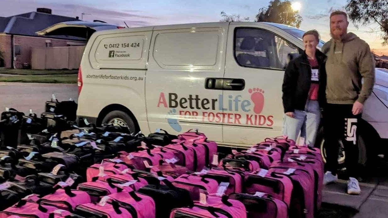 A man and woman stand by a white van with the 'A Better Life for Foster Kids' logo on the side. In front of them are several black and pink suitcases lined up neatly.