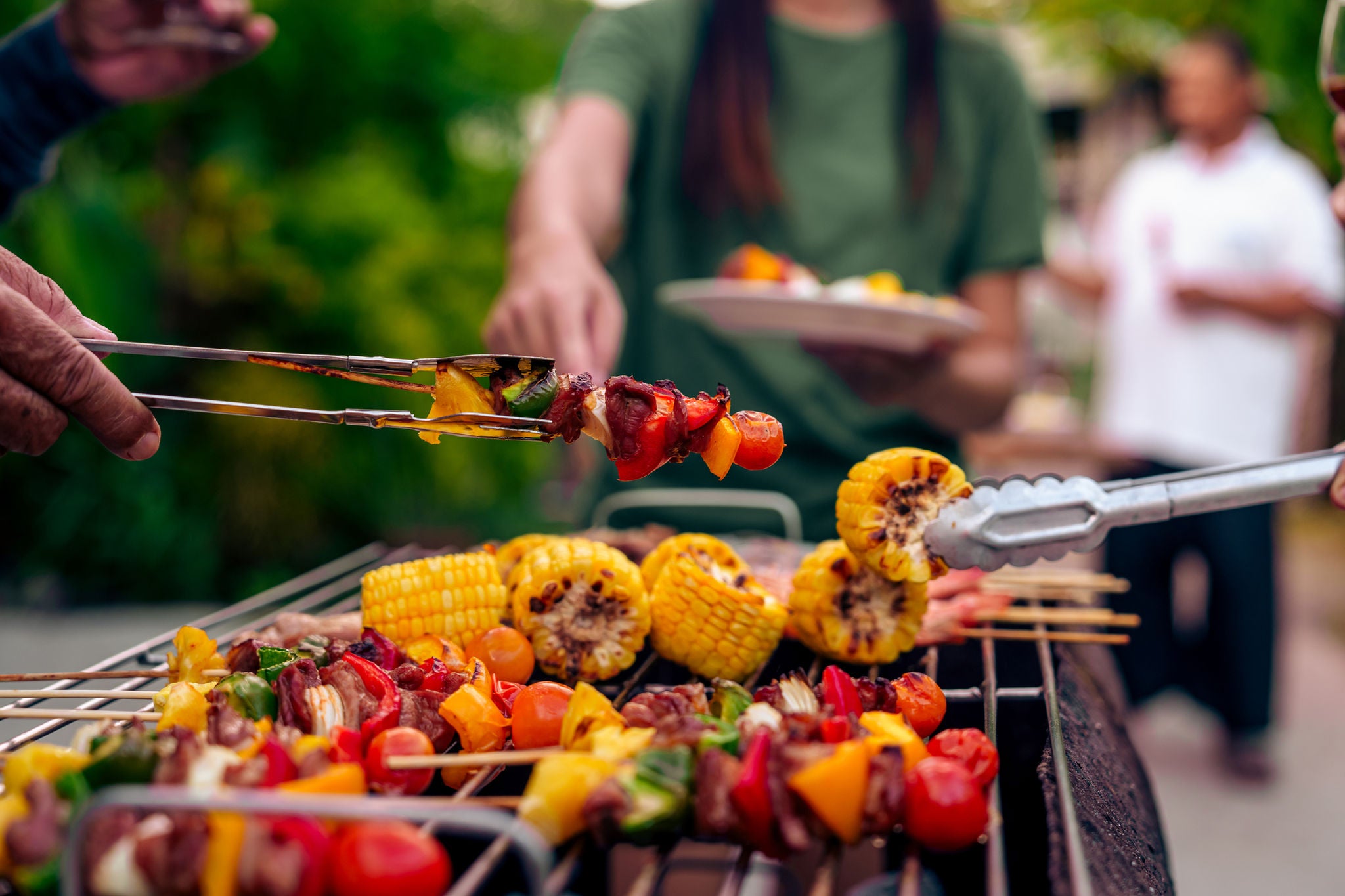 Close up of barbeque showing chargrilled vegetables on skewers and people lifting off the food using tongs
