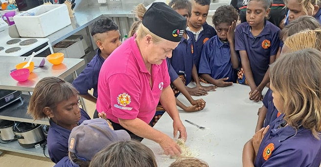 A chef in a bright pink shirt is teaching a group of indigenous children how to make dough