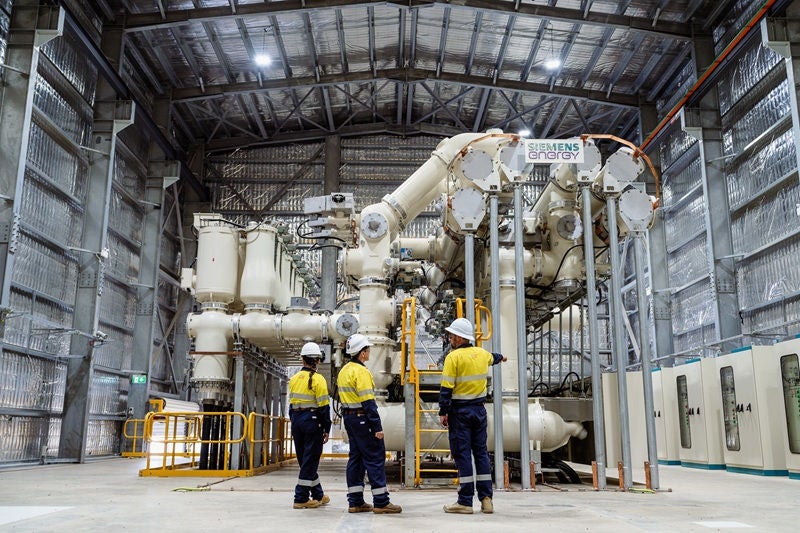 Three workers in high-visibility clothing stand beside large electrical equipment inside an industrial facility.