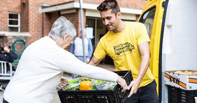 Smiling Oz Harvest worker in a yellow t shirt helping an eldery woman with her food delivery from his van