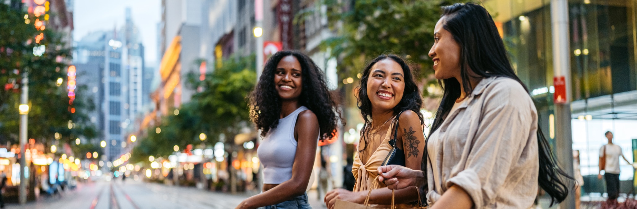 Three smiling women with shopping bags crossing a busy Sydney street at dusk