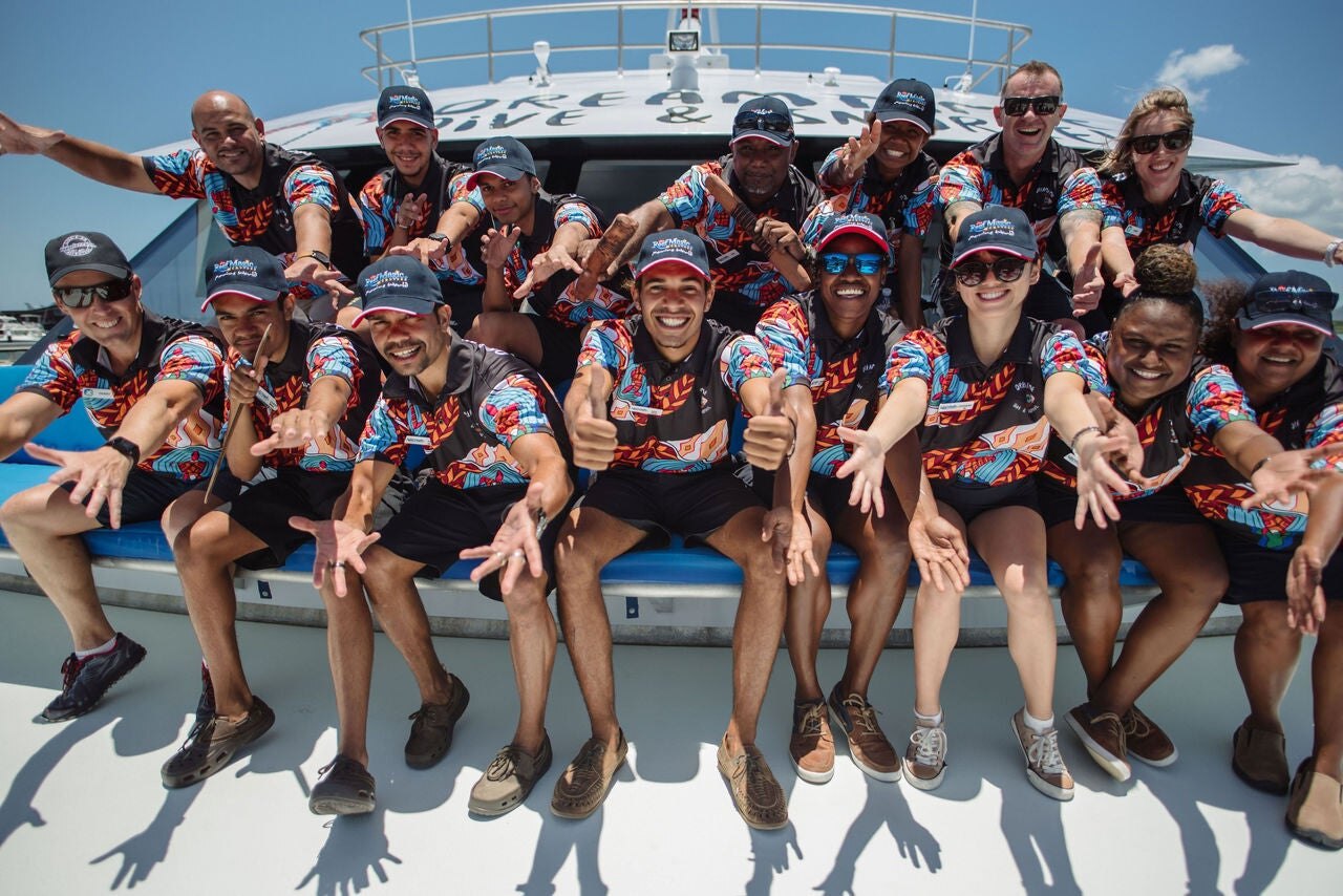 People sitting on the front of a boat smiling with arms outreached. The man in the centre of the group is giving two thumbs up.