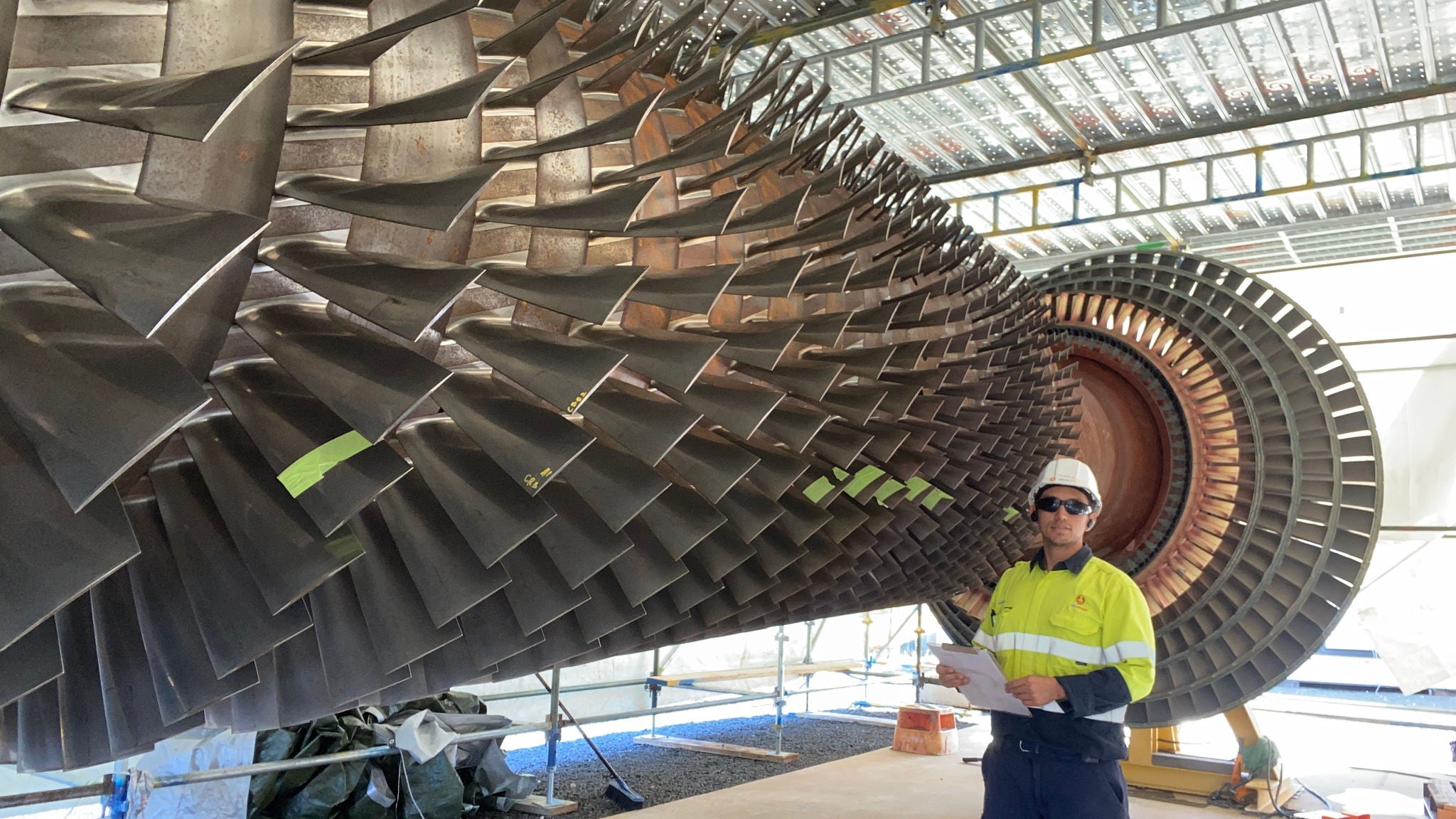 Mario in hi-vis, sunglasses and hard hat in front of an enormous turbine.
