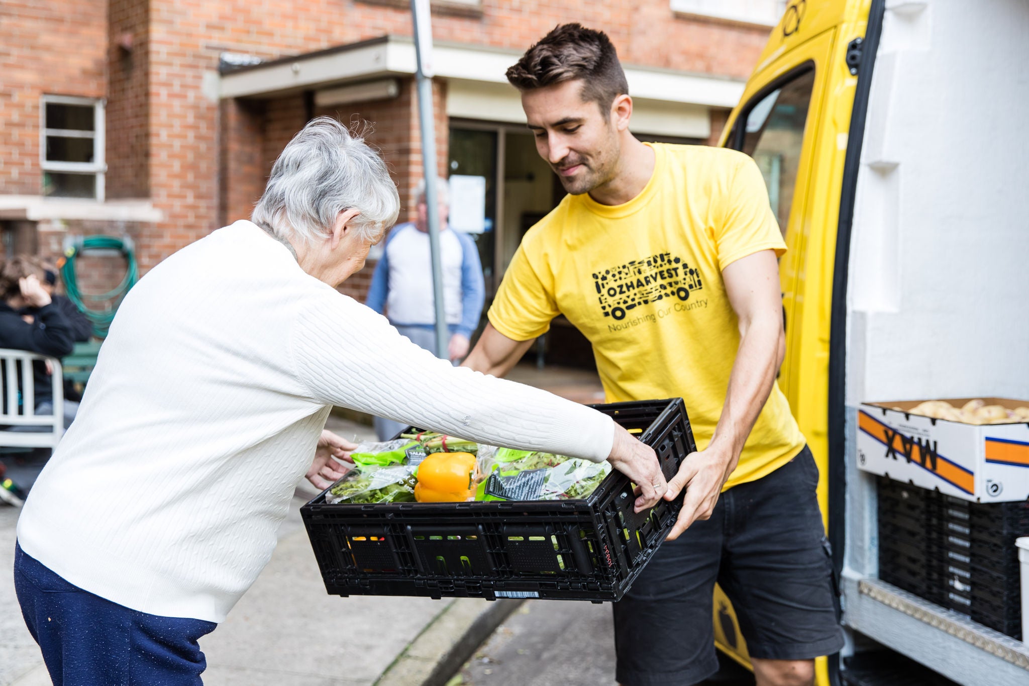 A young man in a yellow OzHarvest t-shirt and dark shorts passes a crate of food from a yellow OzHarvest van to an elderly woman. They are in front of a brick building.