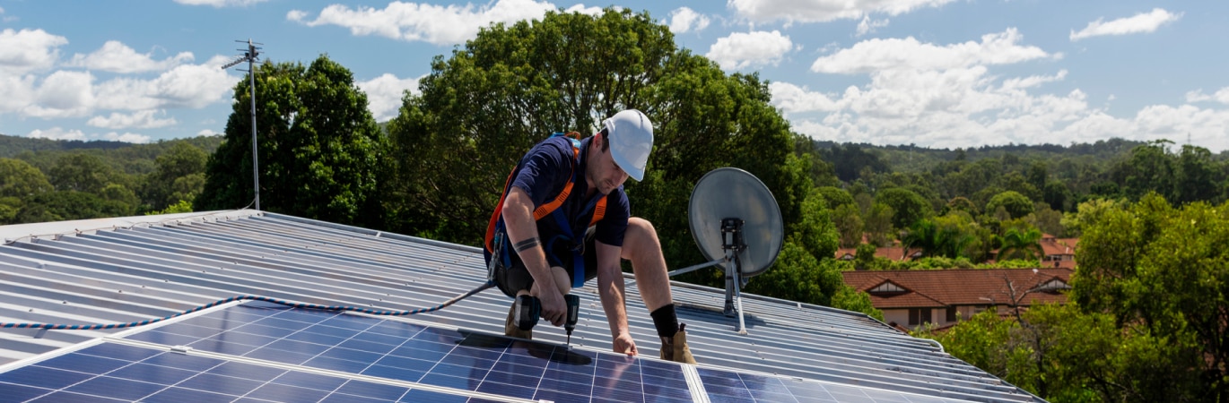 A worker in safety gear in kneeling on a tin roof and affixing solar panels with a drill