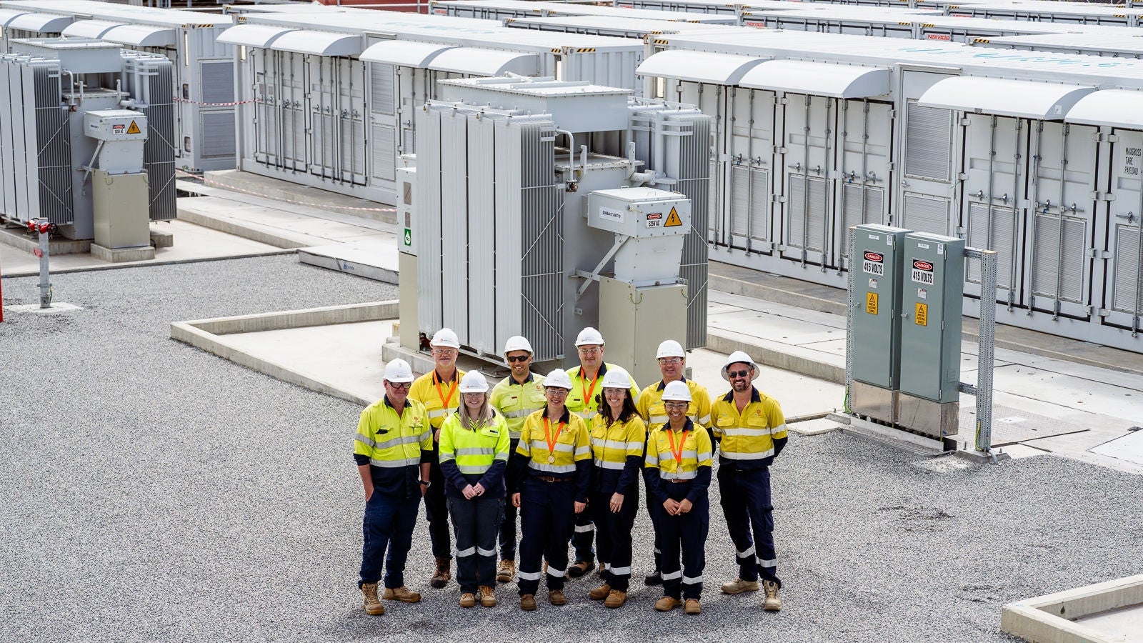 Three workers in high-visibility clothing stand beside large electrical equipment inside an industrial facility.