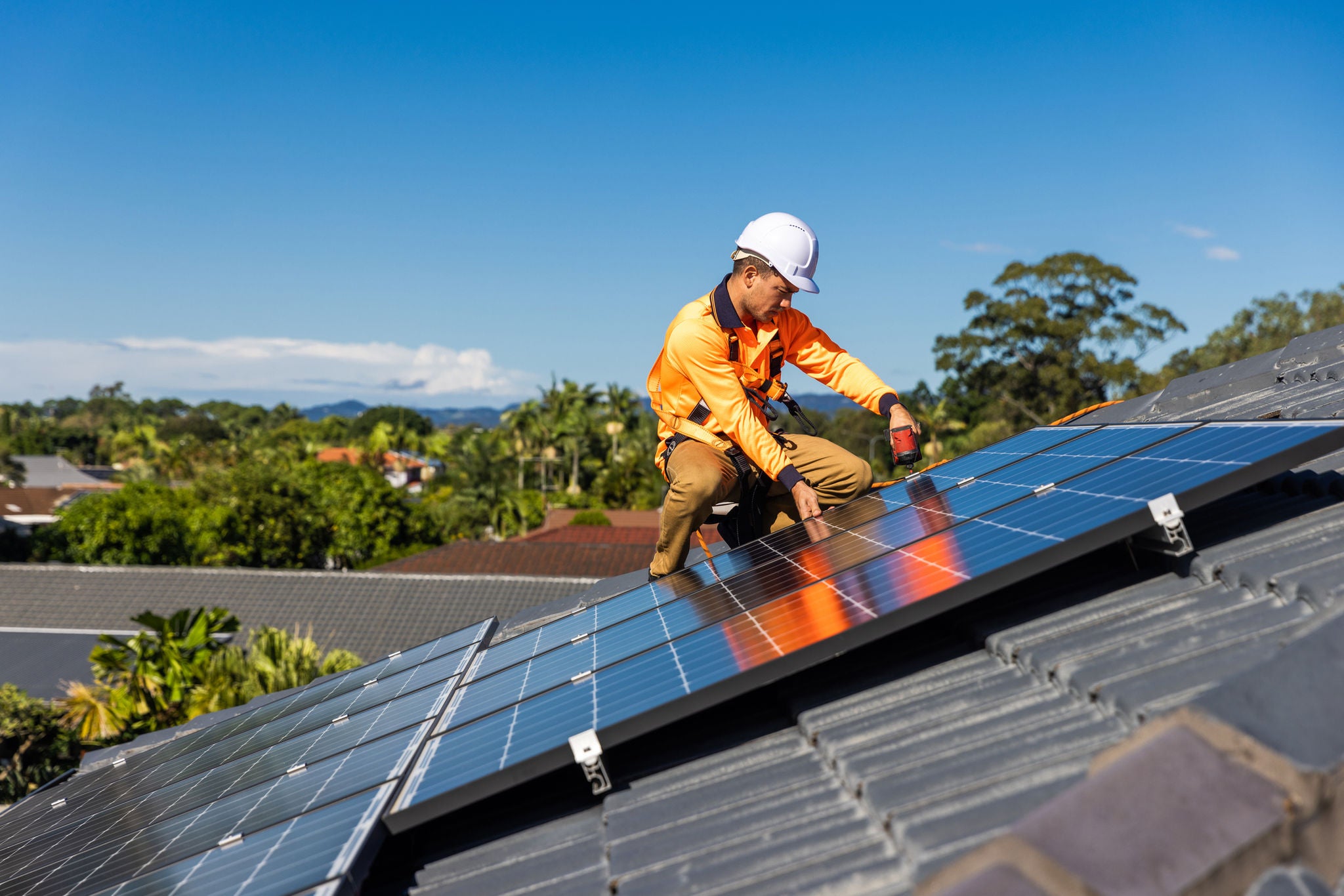 Solar installer in high visibility workwear and hard hat kneeling on a roof and fitting solar panels