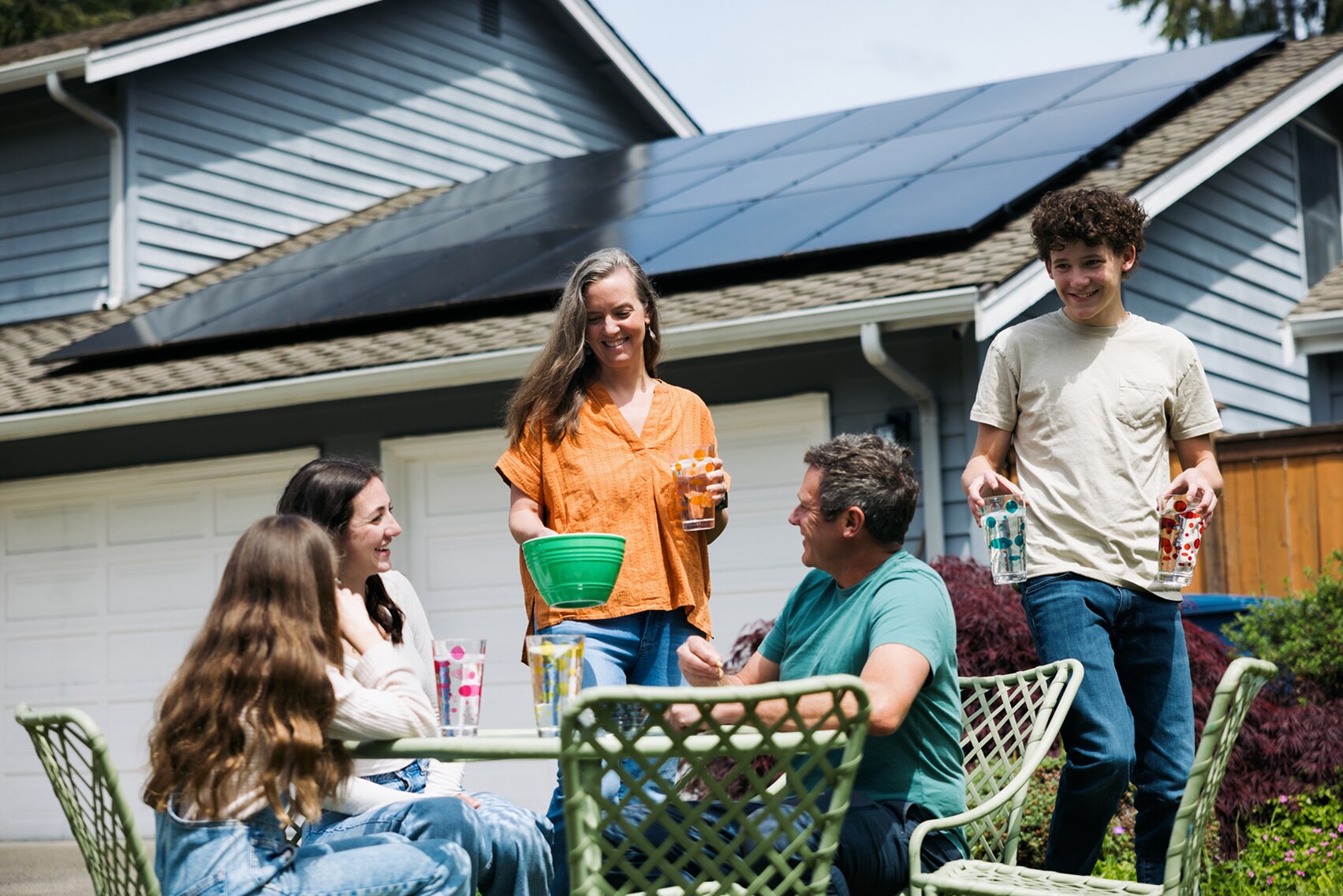 Family smiling around an outdoor table setting with a home in the background showing solar panels on the roof