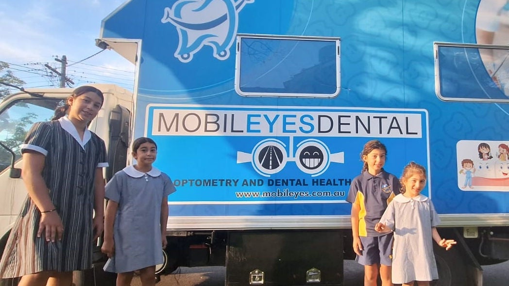 Four young girls stand in front of one of Mobileeyes' blue mobile dental vans.