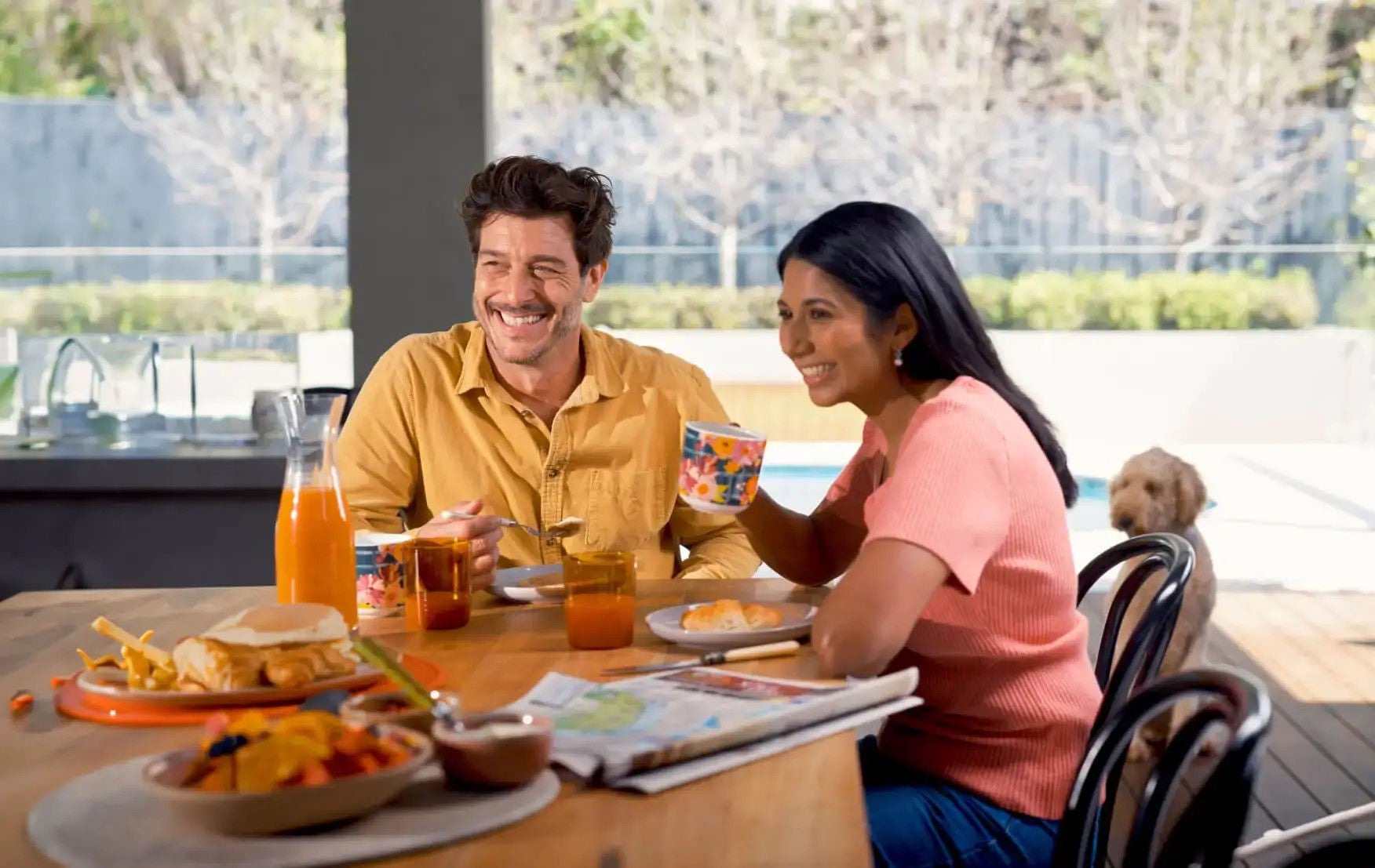 A smiling couple are enjoying breakfast on the deck outside their house while their dog looks on hopefully