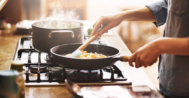 Close up of woman sauteeing vegetables in a fry pan on a gas hob while a large saucepan boils on the next burner