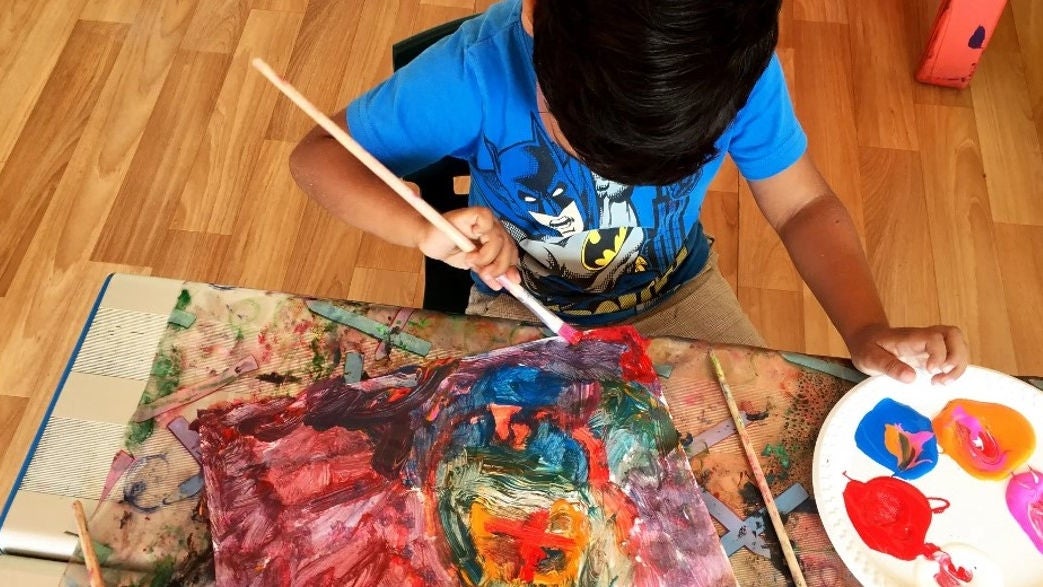 An overhead shot of a child painting at a table. He is wearing a blue Batman t-shirt.