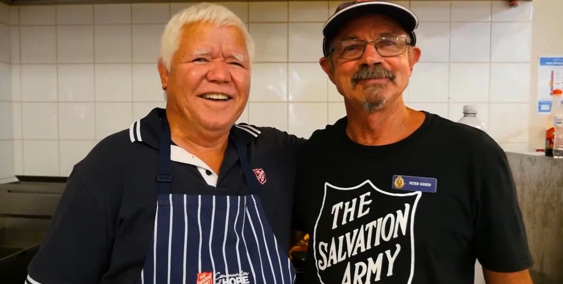 Two Salvos volunteers smile for a picture. The man on the left has white hair and the man on the right wears a cap, glasses and has a moustache and goatee.