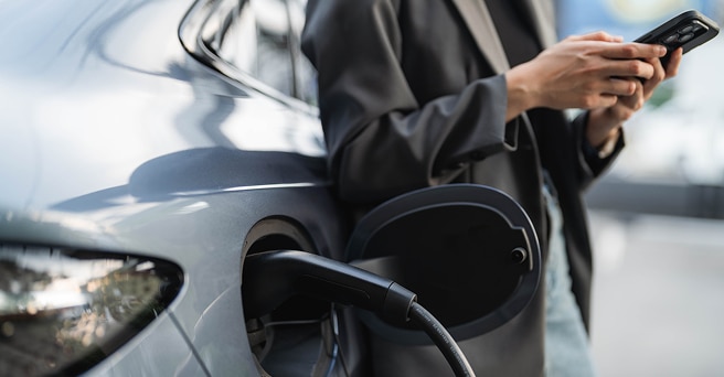 A young woman is using a mobile phone and leaning against an EV that is charging