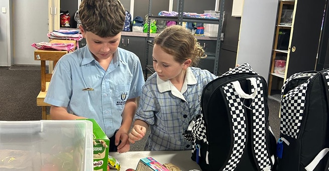 Two schoolchildren packing pantry items into black and white backpacks for charity