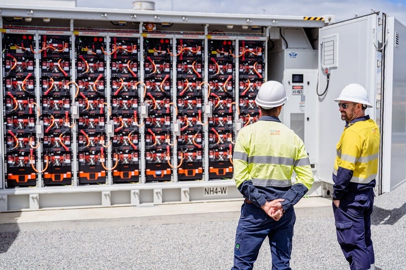 Two workers in high-visibility clothing and hard hats stand beside an open battery storage unit.