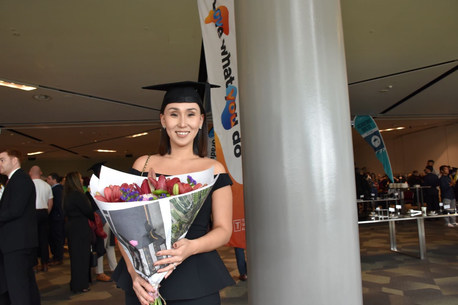 An image of Mika during her graduation. She is smiling, holding a large bouquet of flowers, wears a black dress and mortarboard cap.