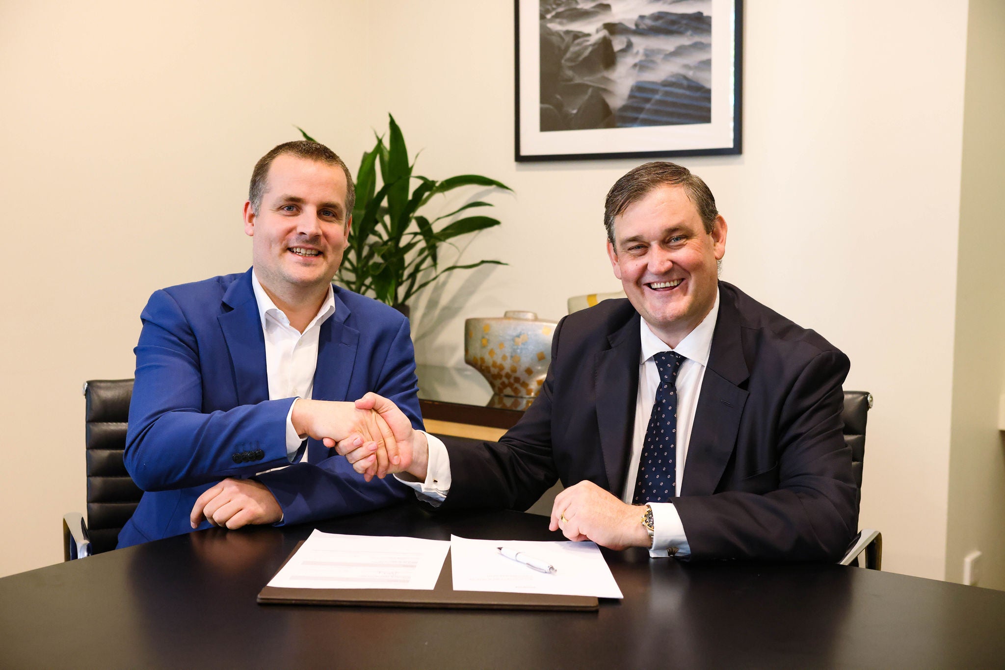 Parkwind Head of M&A Mathias Van Steenwinkel, wearing a blue suit, shakes hands with Alinta Energy MD&CEO Jeff Dimery as they sign their agreement. Both men are smiling. 