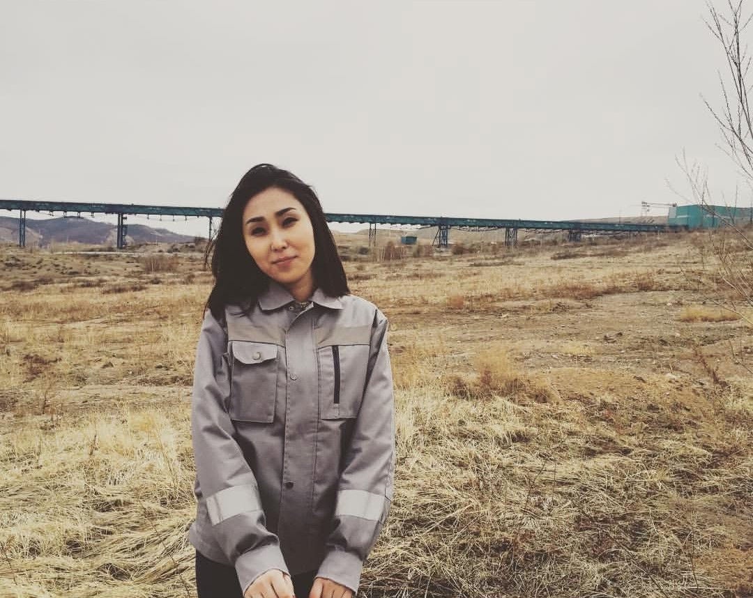 Mika stands in a dry, grassy field wearing a grey jacket with reflective stripes. Behind her, an industrial structure—possibly a conveyor belt or bridge—stretches across the landscape under an overcast sky, with distant hills in the background.