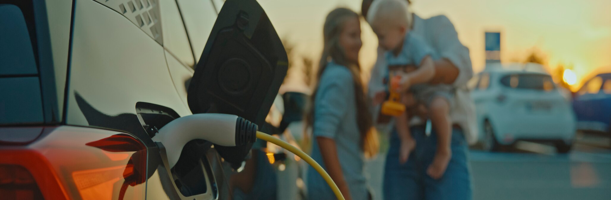 A close shot of an EV charging with a young family in the background at sunset
