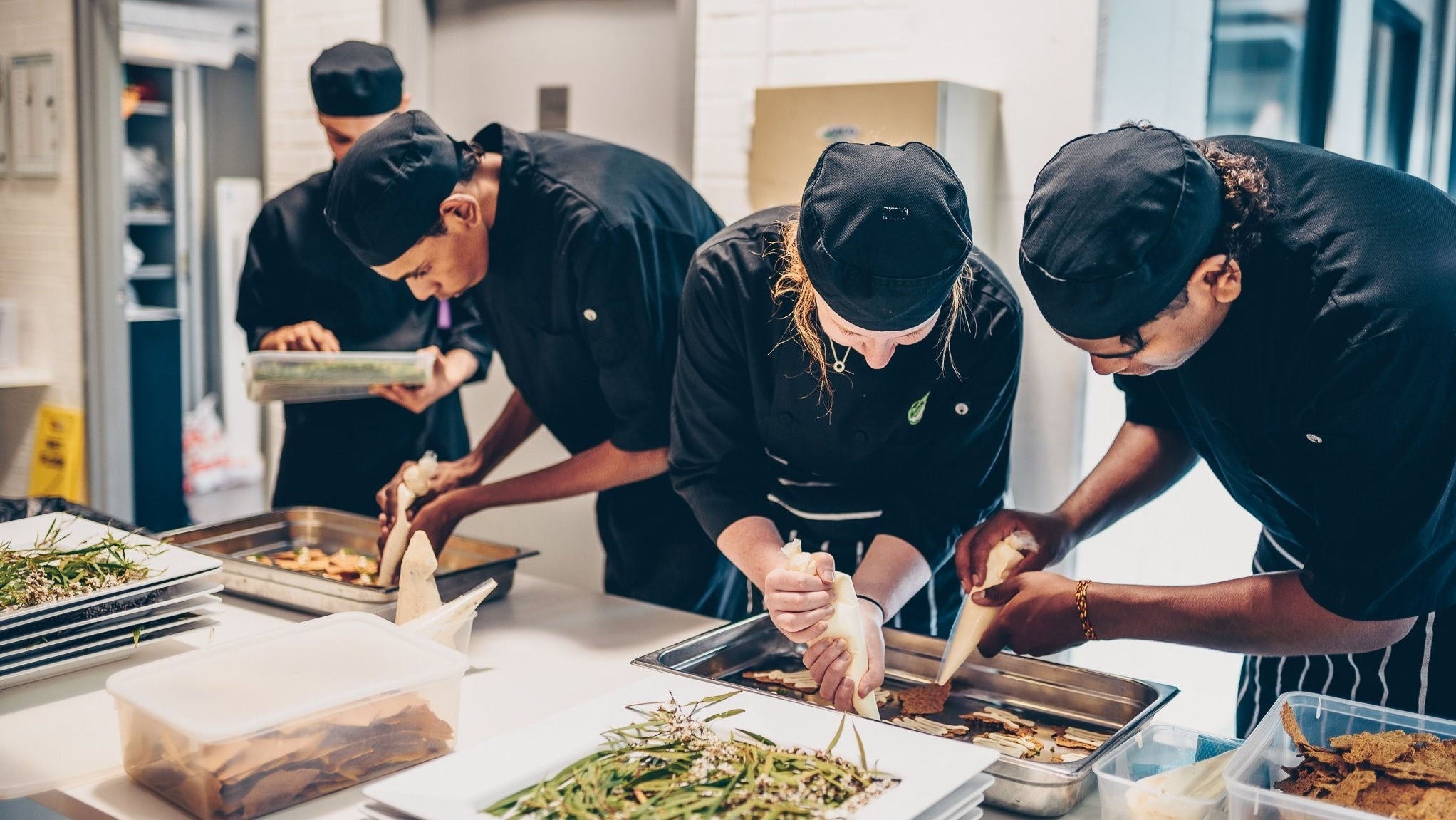 Four chefs in black clothes and hats prepare canapes in a kitchen.