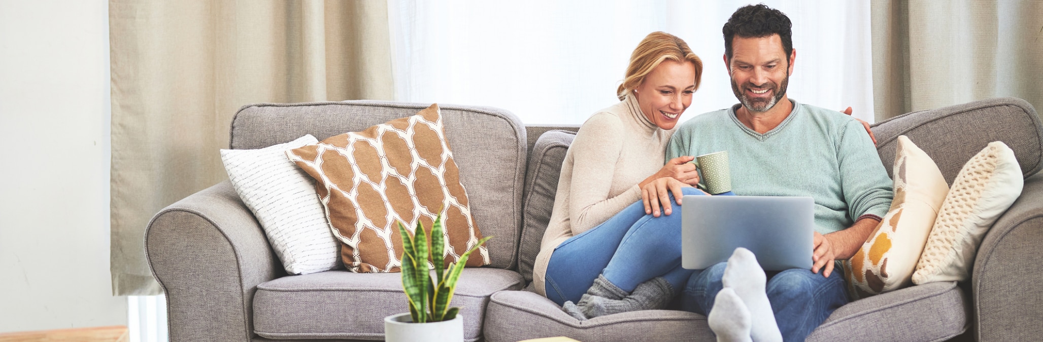 A smiling couple a sitting close together on a sofa with a cup of tea and looking at a laptop screen in a light airy lounge room