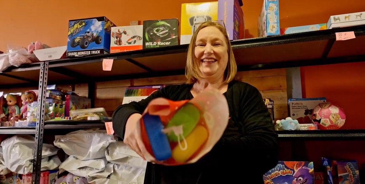 A smiling blonde woman holding a colourful toy wrapped in gift wrapping. She stands in front of shelves filled with boxed toys and dolls.