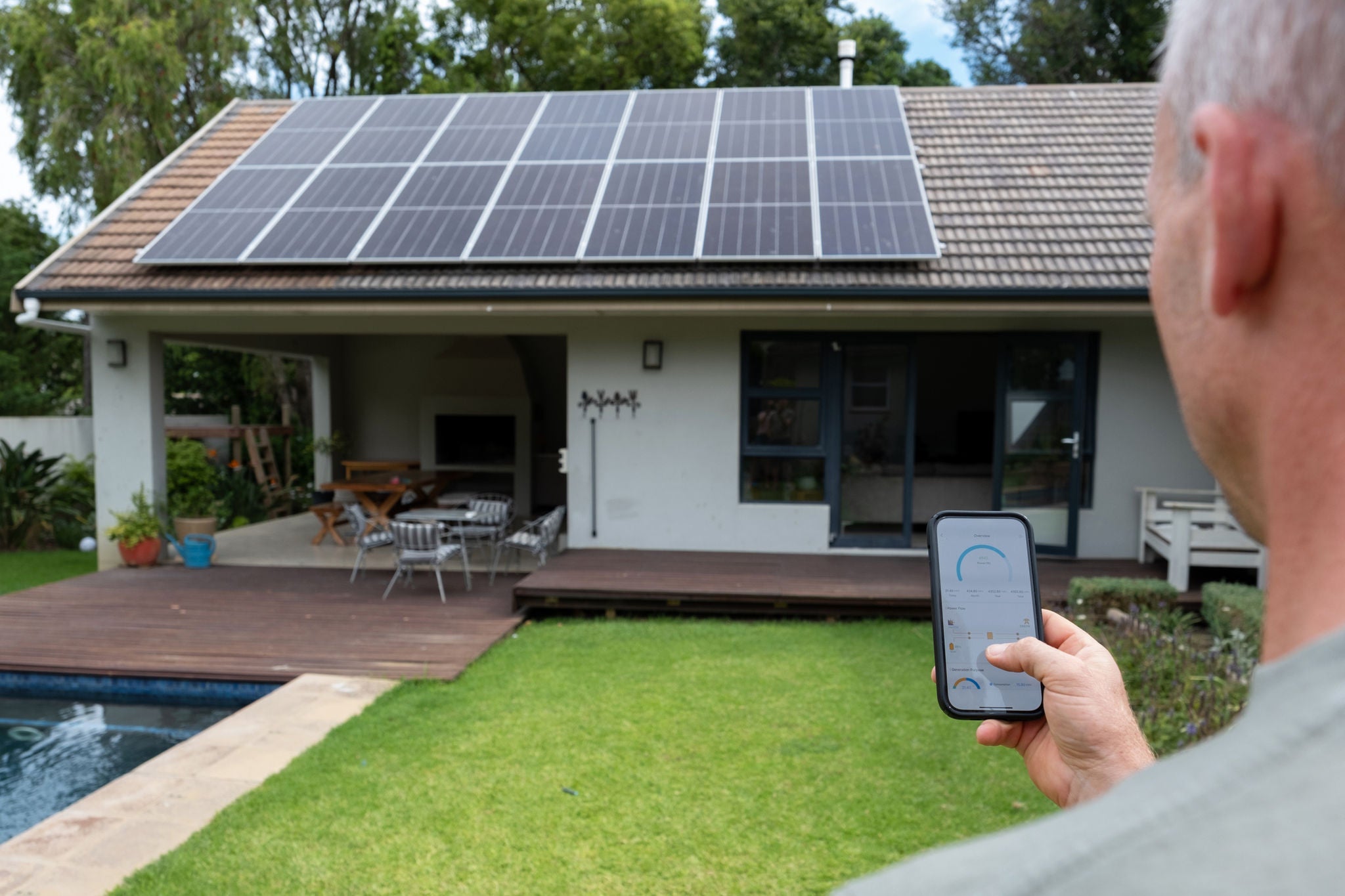 Man holding mobile phone with application on the screen and looking at the back of his house roof which is covered with solar panels