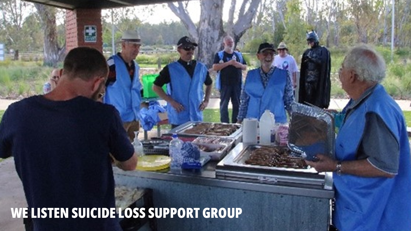 Several people attend a public barbecue in a park-type setting where they are cooking sausages. Most of them are wearing blue vests. One of the men, seen in the background, is dressed as Batman.
