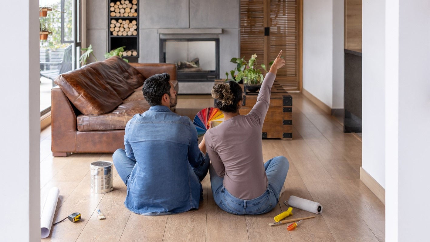 View of couple from behind, sitting on a wooden floor and choosing colours from a paint swatch guide. The woman is pointing up at a wall.