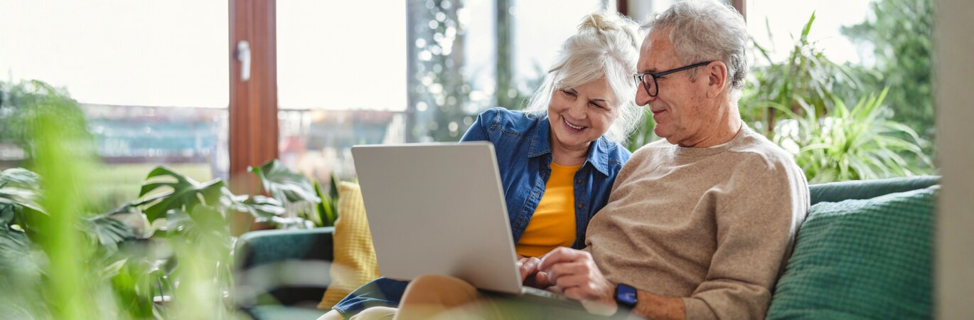 A mature couple are viewing a laptop and sitting on a green sofa in a bright and airy room surrounded by plants