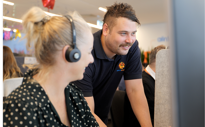 A smiling blonde woman wearing a headset is sat next to a standing, smiling man with short hair, a moustache and an Alinta Energy polo top in an office environment.