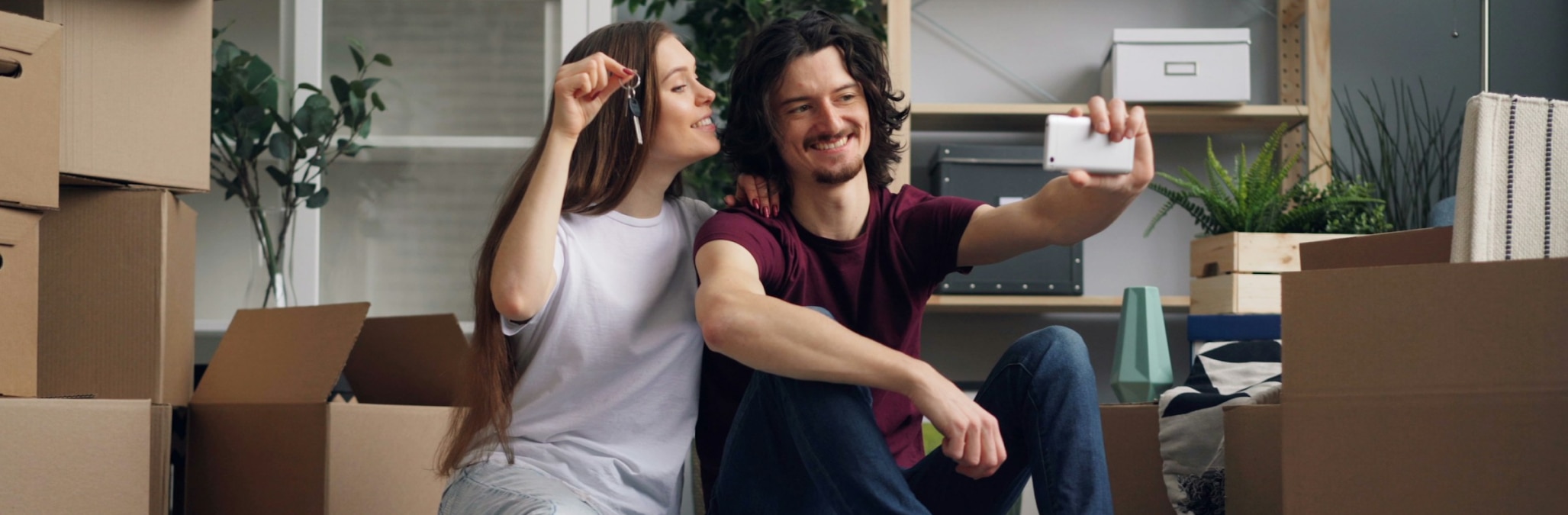 A couple sitting on a floor surrounded by moving boxes but taking a selfie and holding up keys to a new house
