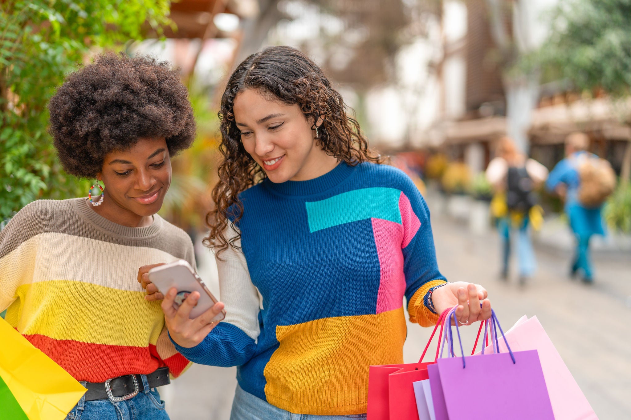 Two smiling women looking at a mobile phone and holding very colourful shopping bags in street filled with greenery and flowers.