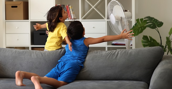 Two children on a sofa spreading their arms in front of a fan on a hot day