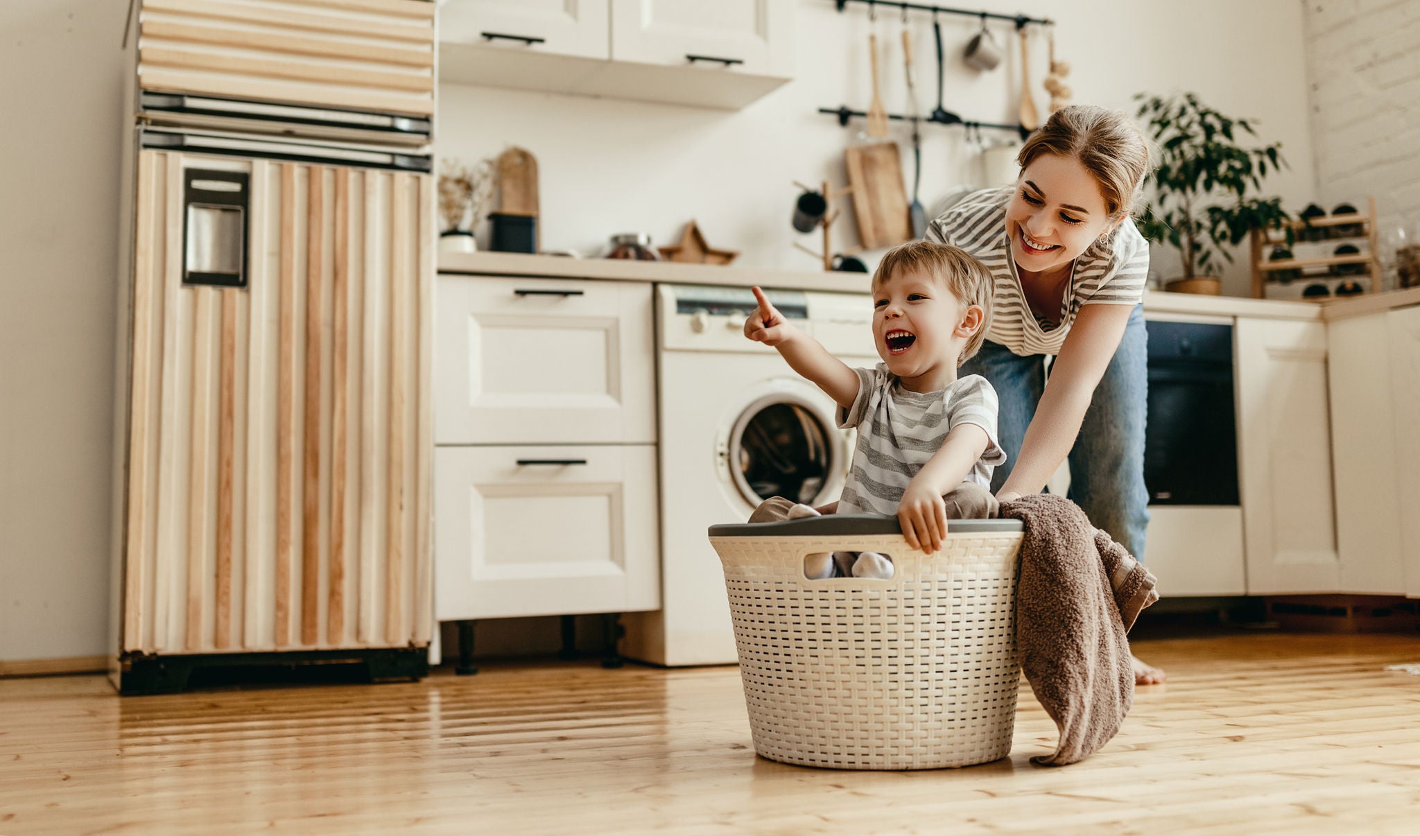 Smiling mother pushing a young boy in a laundry basket across the floor of a light and modern kitchen