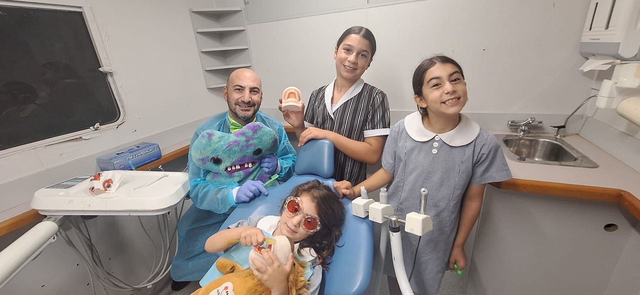 A male dentist smiling with three young girls. One of them is in the dentist chair wearing eye protection.