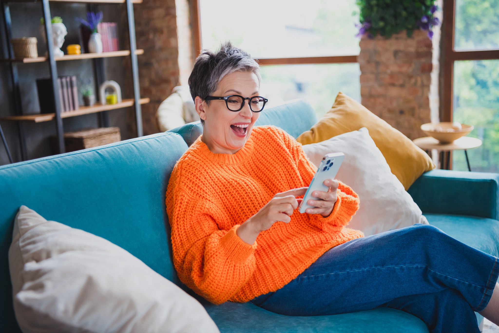 Smiling mature woman in bright orange sweater browsing her mobile phone while sitting on a blue sofa