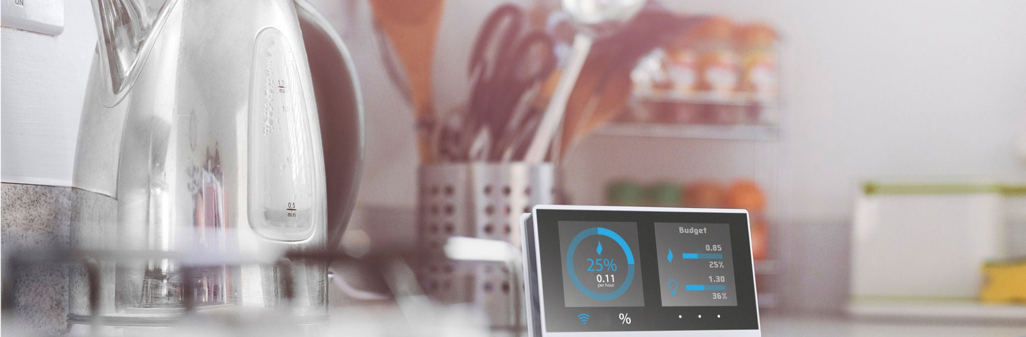 A close up of a smart device showing current energy use on its display next to a chrome kettle in a bright kitchen