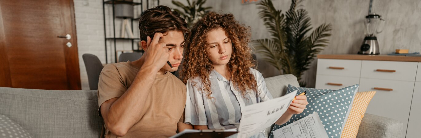 A young couple are sitting on a sofa and reading through bills together
