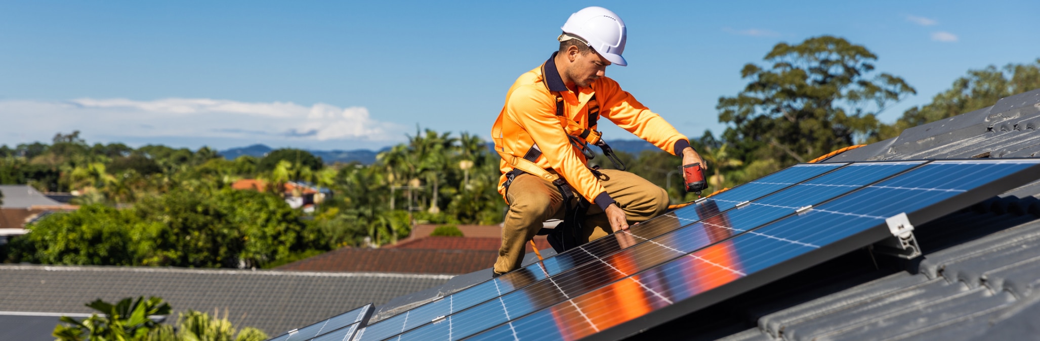 Solar installer in high visibility workwear and hard hat kneeling on a roof and fitting solar panels